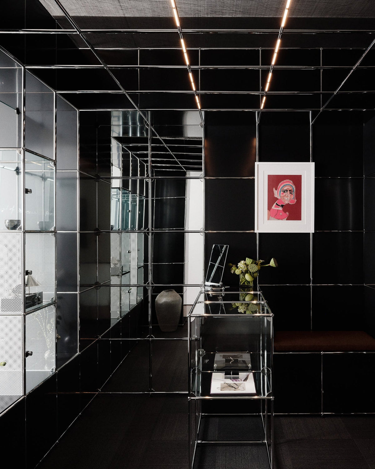 A modern interior space with glossy black mirrored tile walls and floor, a small glass table with flowers, built-in shelving, and a framed red artwork hanging on the back wall.