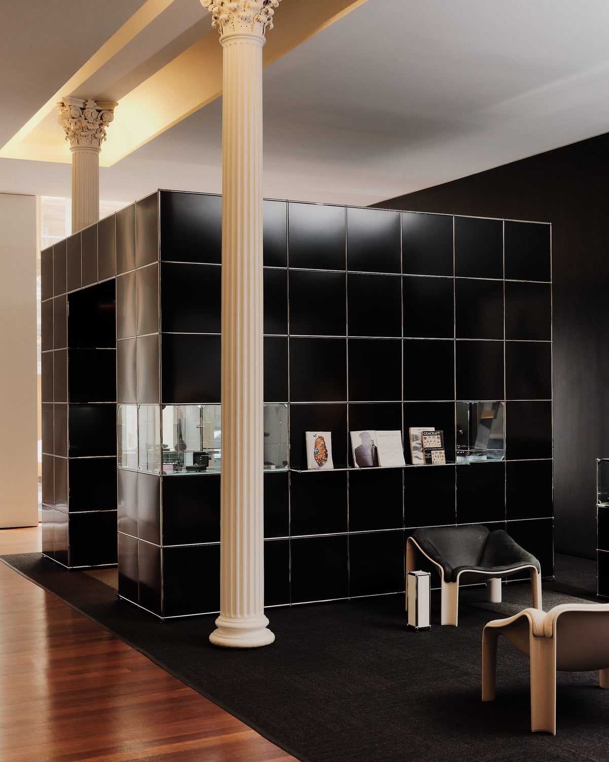 A modern black modular wall display or partition made of glossy square panels, set in a stylish interior with white fluted columns, dark wood flooring, and minimalist seating.