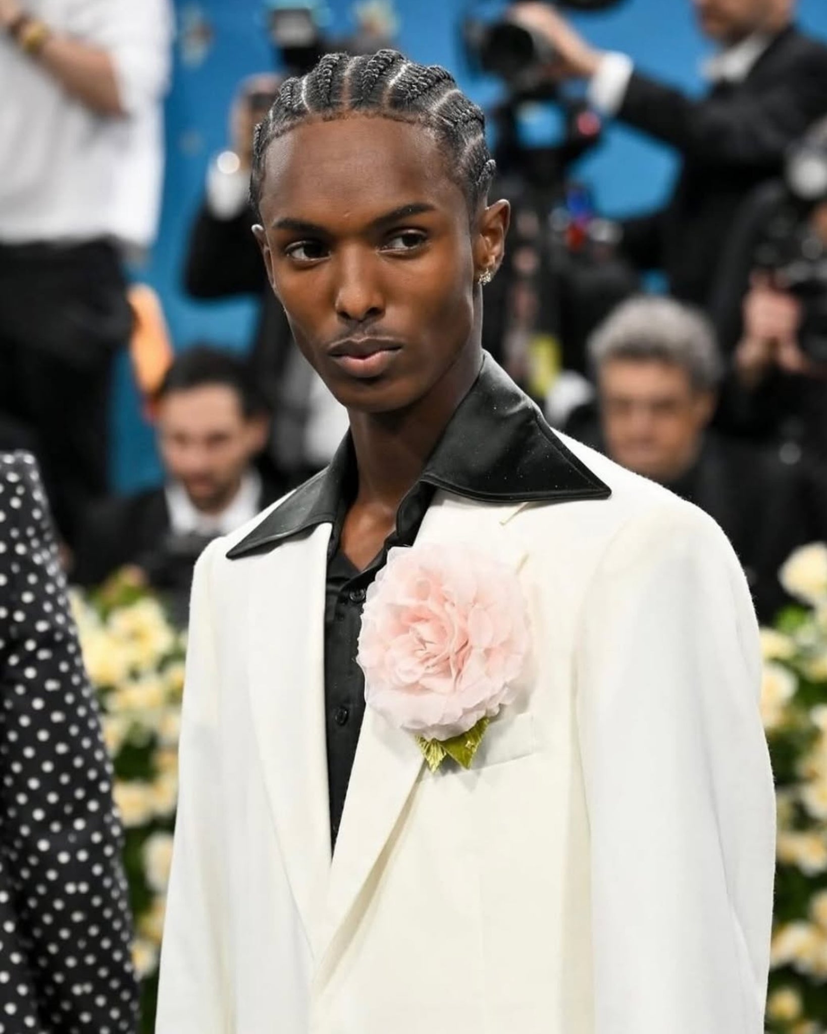 Model on a fashion runway wearing a white coat with a pink flower, blurred audience in the background