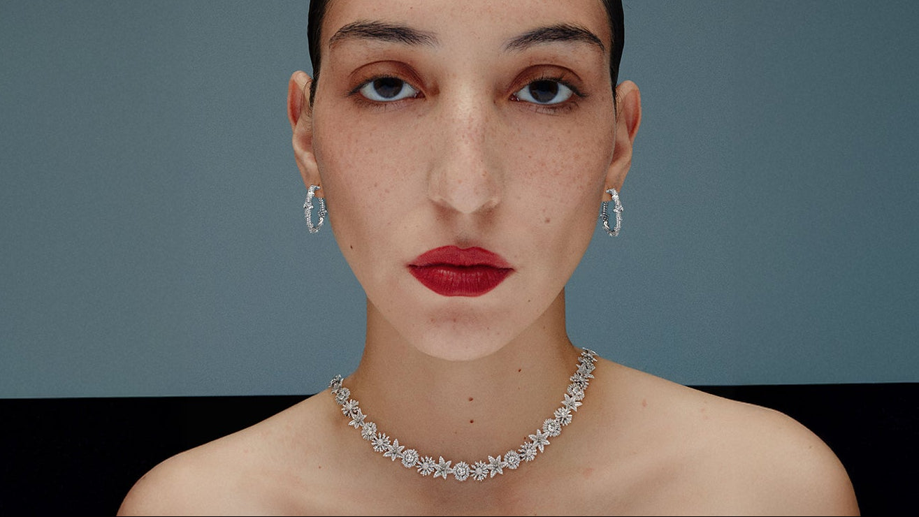A close-up portrait of a model wearing a sparkling diamond necklace and matching diamond drop earrings against a dark gray background.